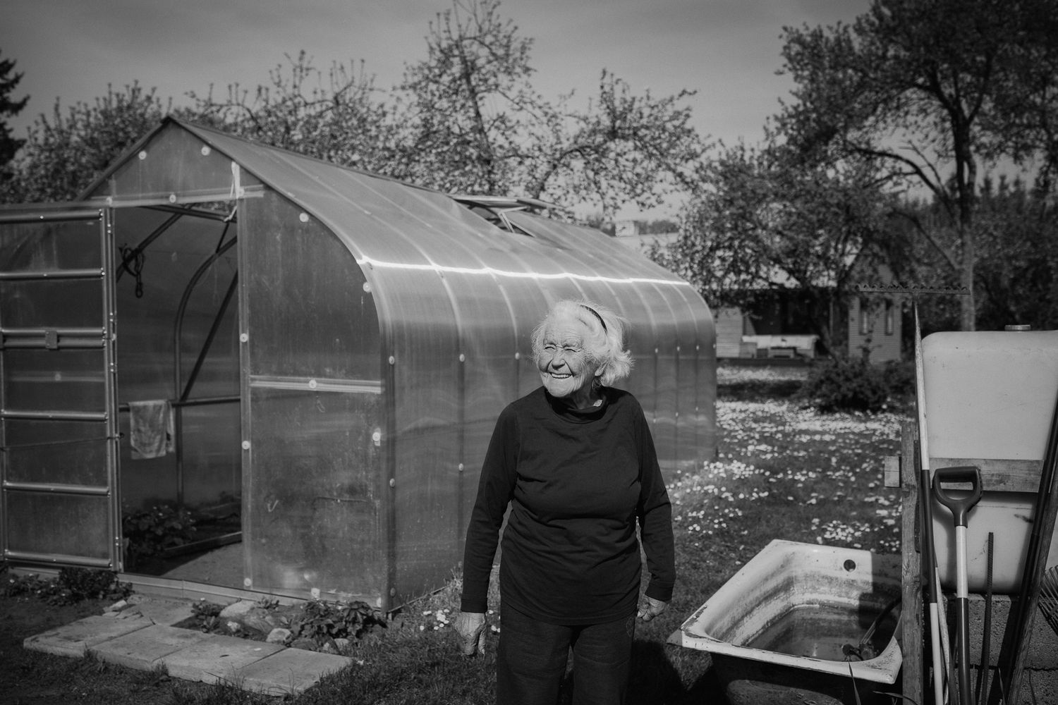 A black and white photograph shows a figure in dark clothing standing beside a curved metal greenhouse in a garden setting.