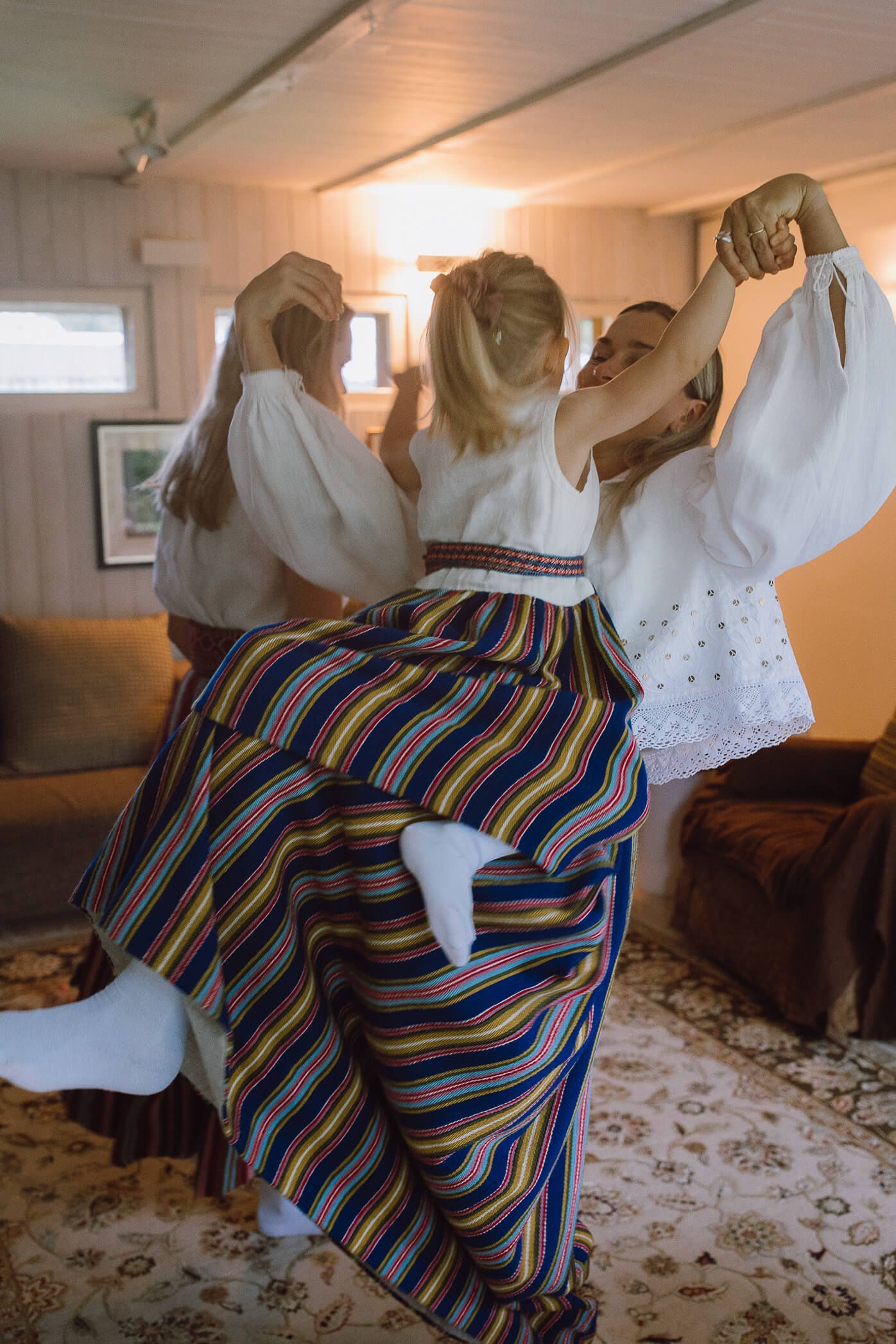 Women in traditional Nordic folk costumes dancing joyfully in a cozy living room setting.