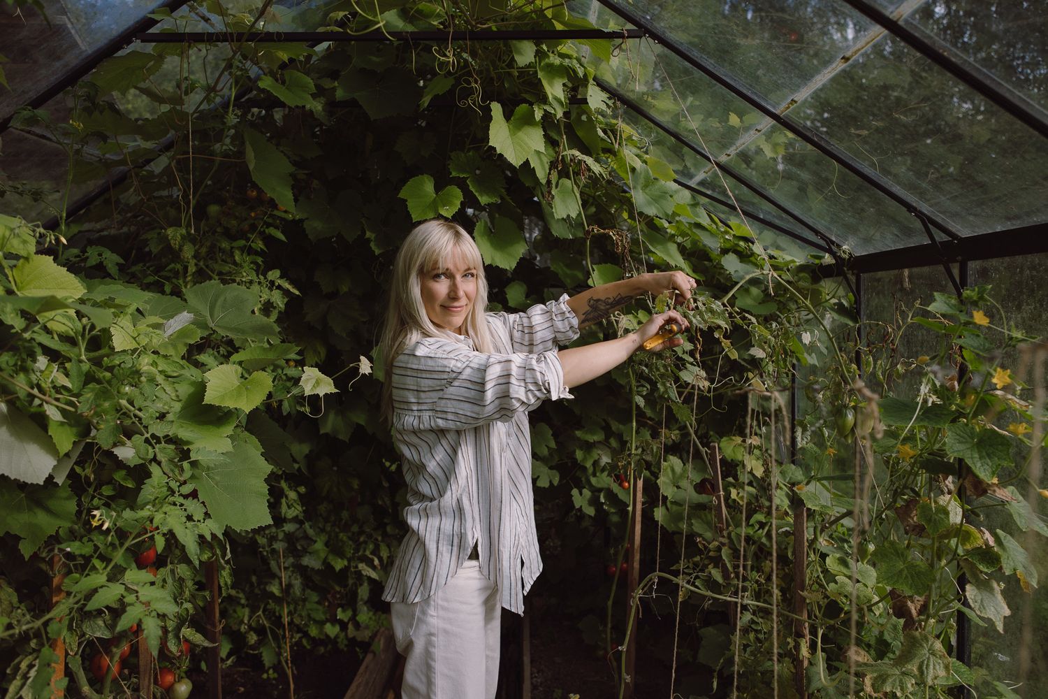 Woman in white clothing tending to plants in a lush greenhouse setting.