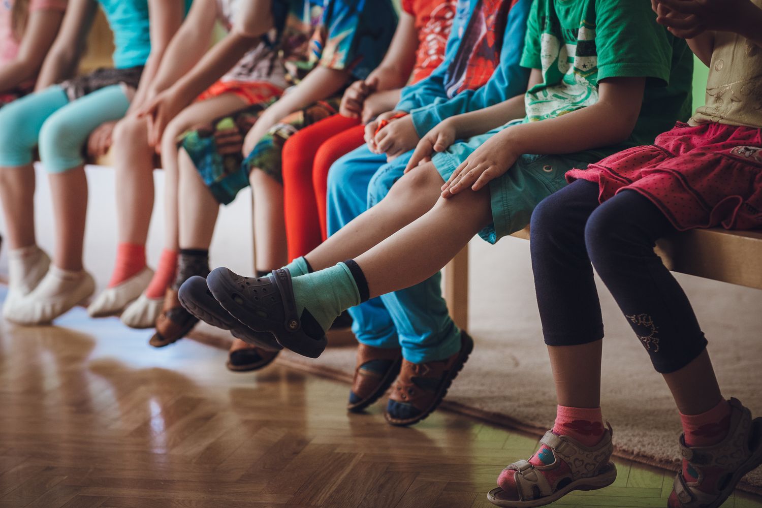 Row of children's feet and legs sitting on benches in an indoor space.