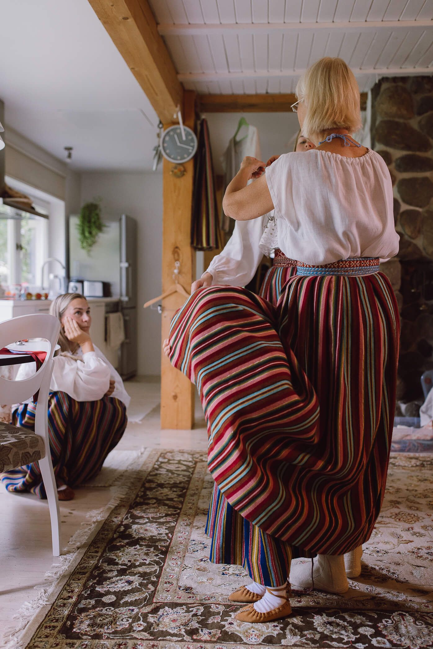 Two women in traditional striped folk costumes and white blouses inside a rustic home interior.