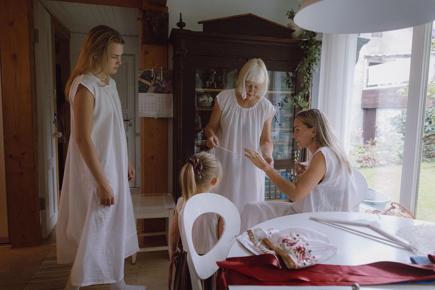 Three generations of women in white dresses gathered around girl at dining table.