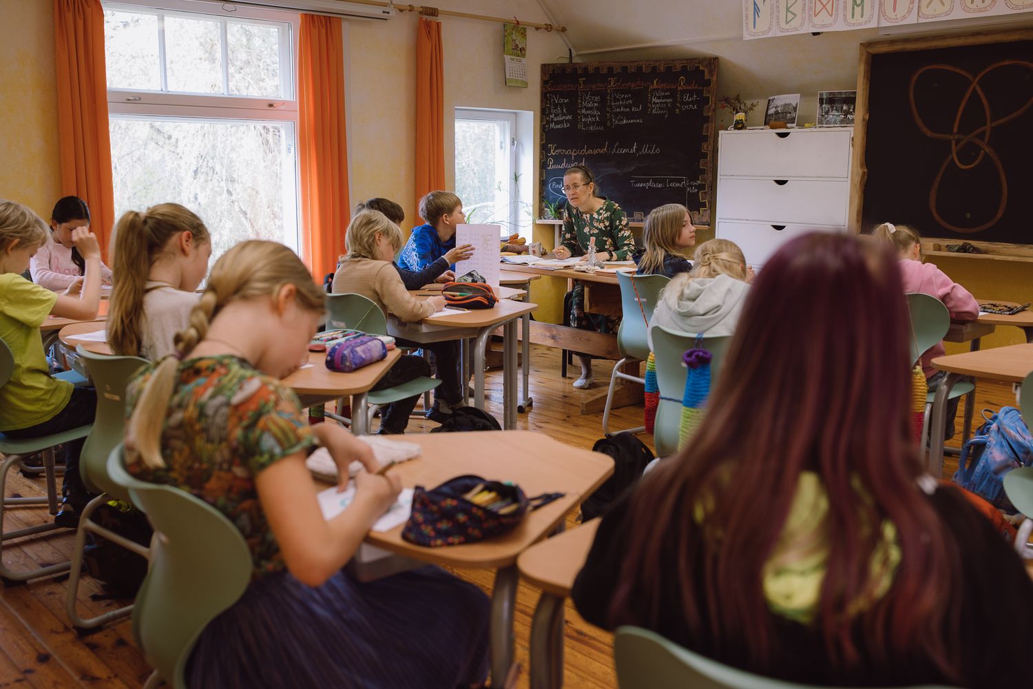 Students participate in classroom activities at wooden desks with orange curtains in background.