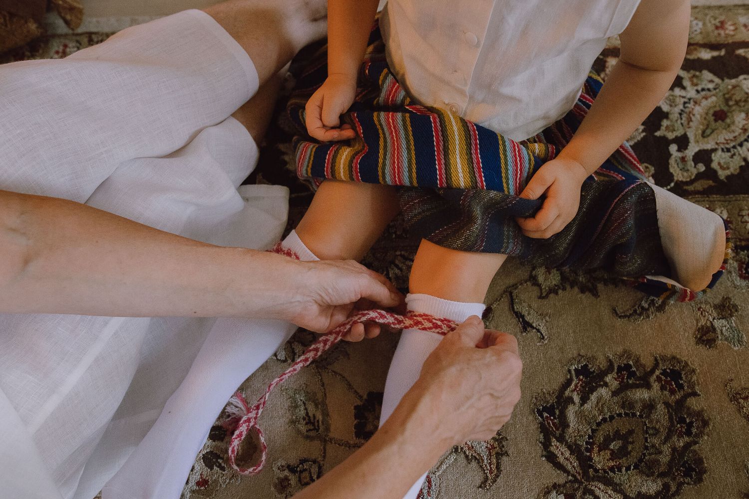 Adult hands tying red and white laces on a child's white sock while the child sits on a patterned rug.