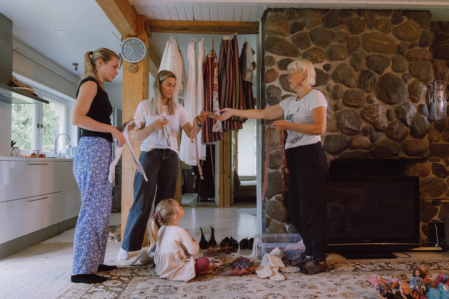Three women and a young girl gather around a stone fireplace in a cozy home, sharing items on a patterned rug.