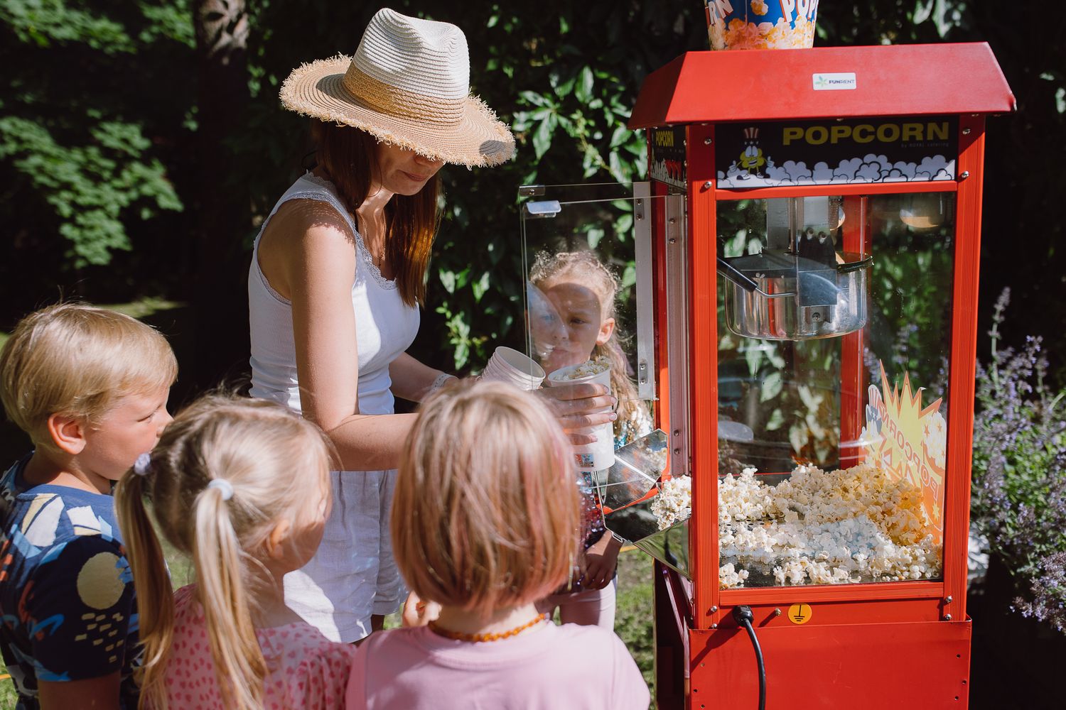Children gather around a popcorn machine with an adult wearing a sun hat.