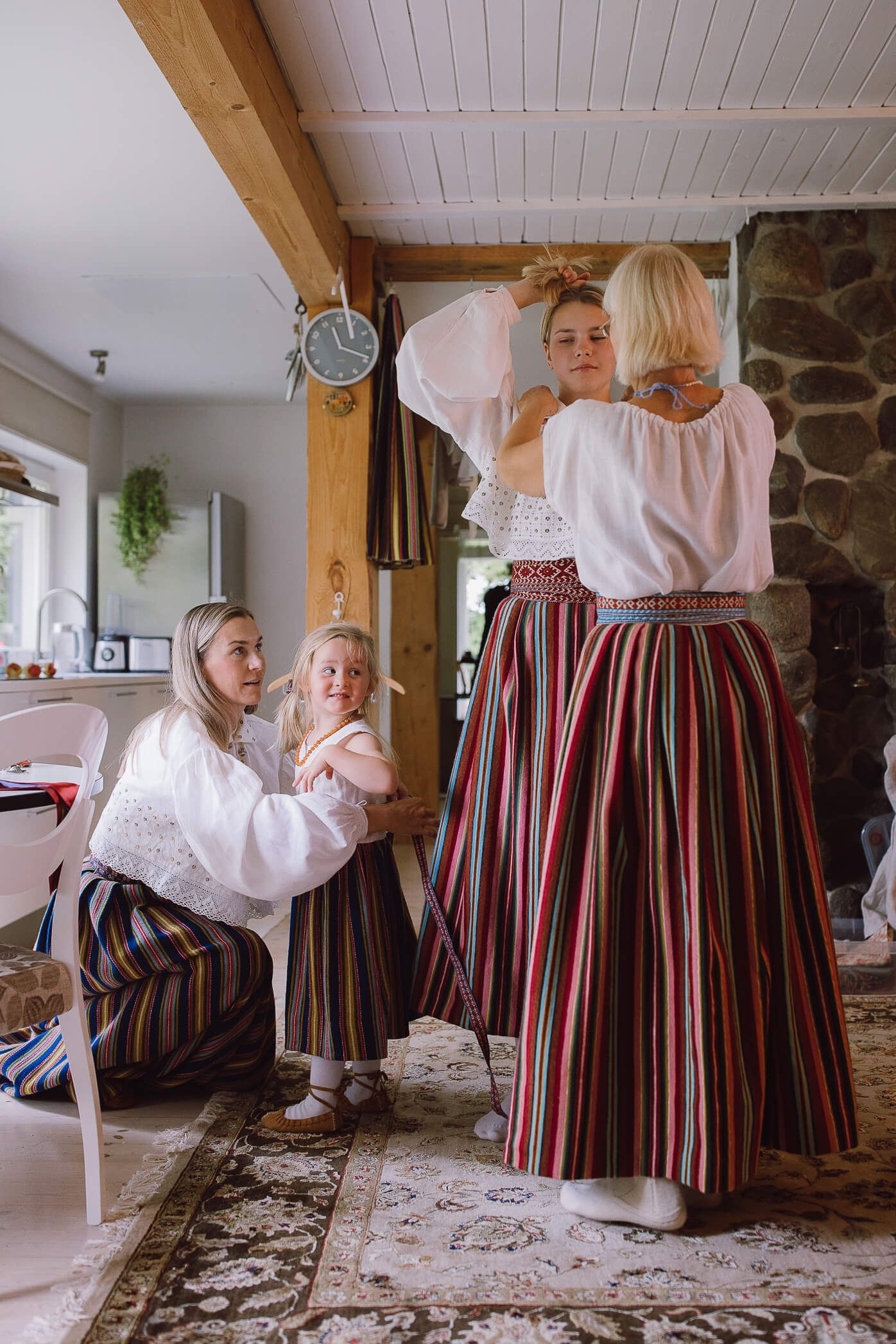 Women and child in traditional striped folk costumes preparing indoors near stone fireplace.
