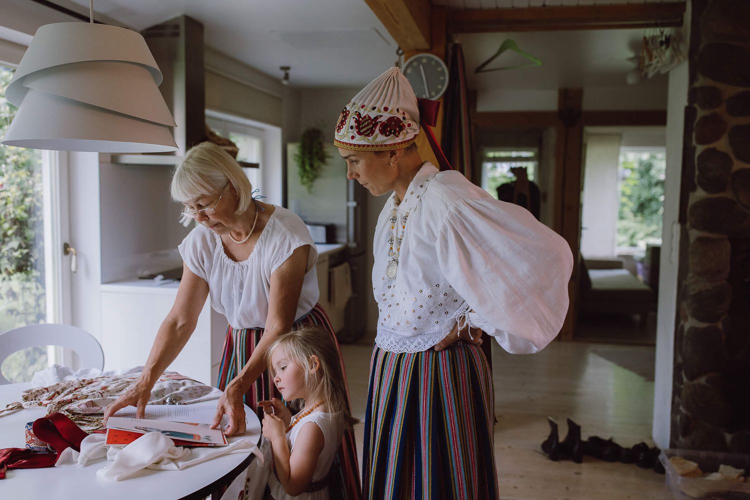 Two women and a young girl in traditional folk costumes prepare items on a table inside a rustic home.