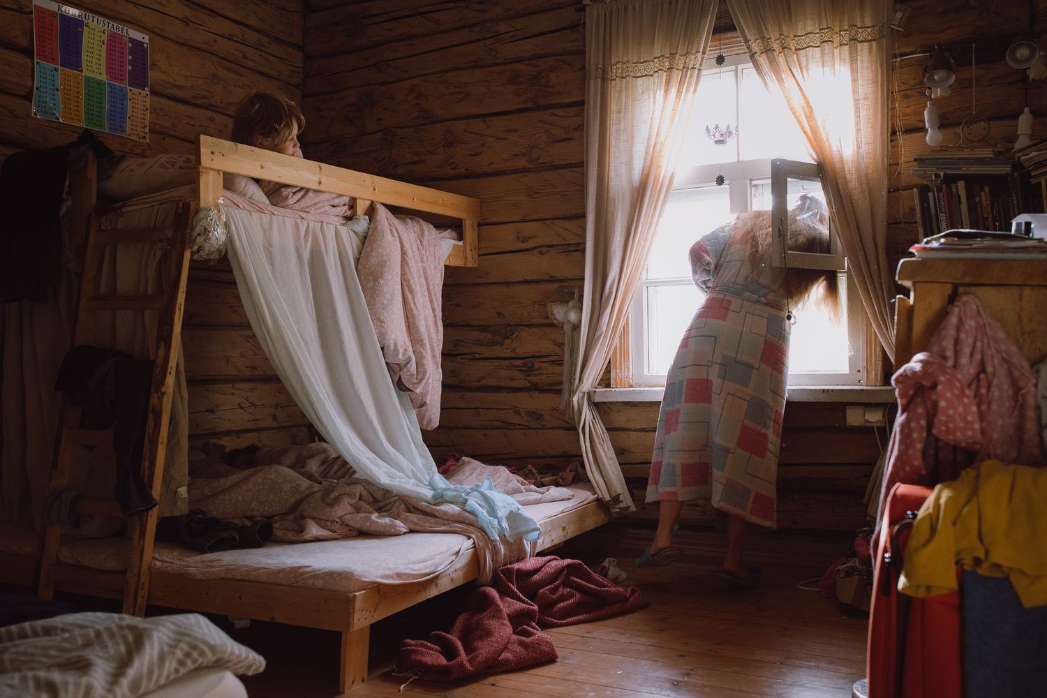 Rustic log cabin interior with wooden walls and white curtains filtering sunlight.