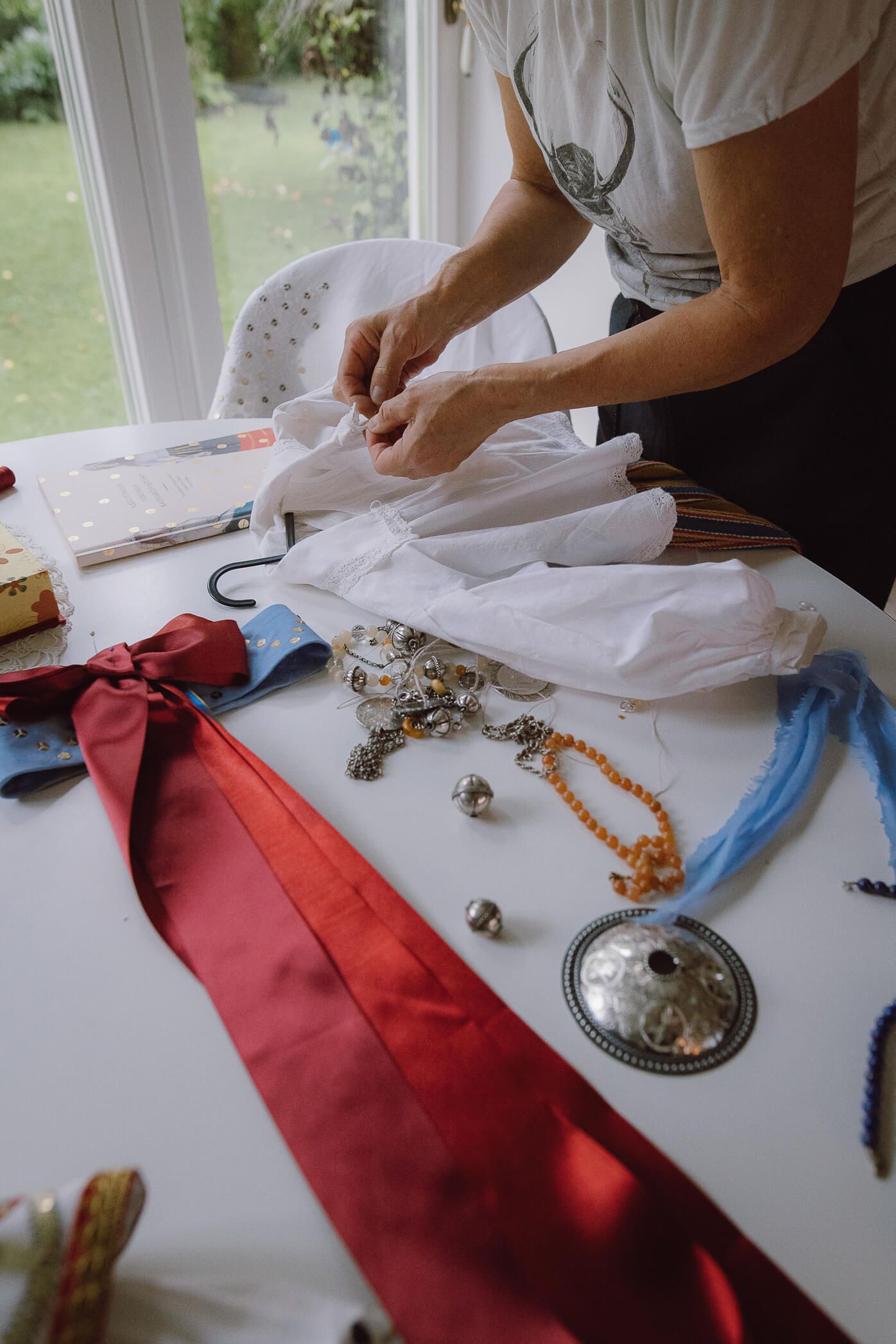 Woman preparing traditional folk costume accessories including ribbons, jewelry, and beads on a white table.