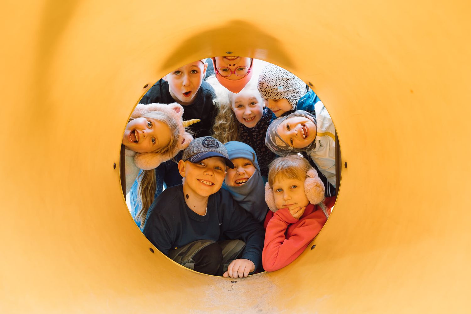 A series of circular cutouts in an orange wall reveal groups of people peering through from the other side.
