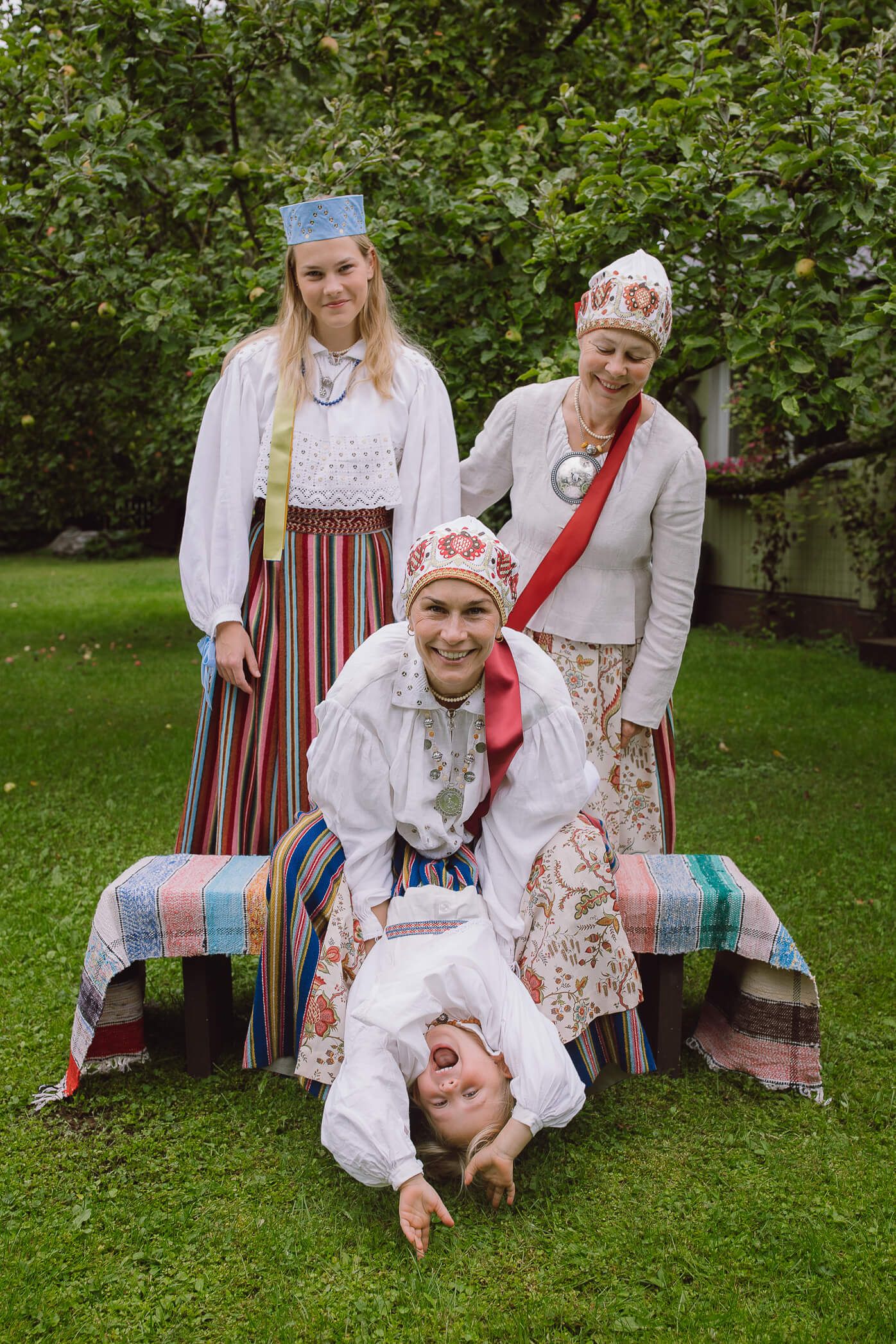 Four women in traditional Estonian folk costumes pose playfully outdoors, one doing a headstand on a colorful bench.