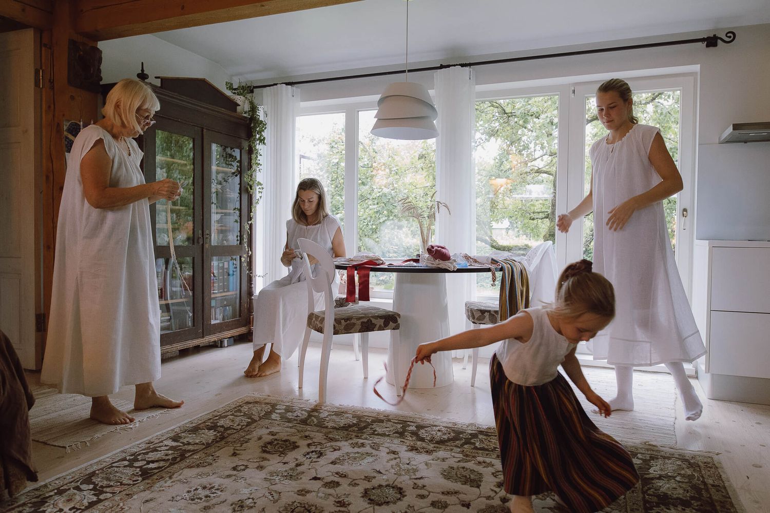 Four women and a girl in white dresses gather in a bright Scandinavian living room with wooden furniture and garden views.