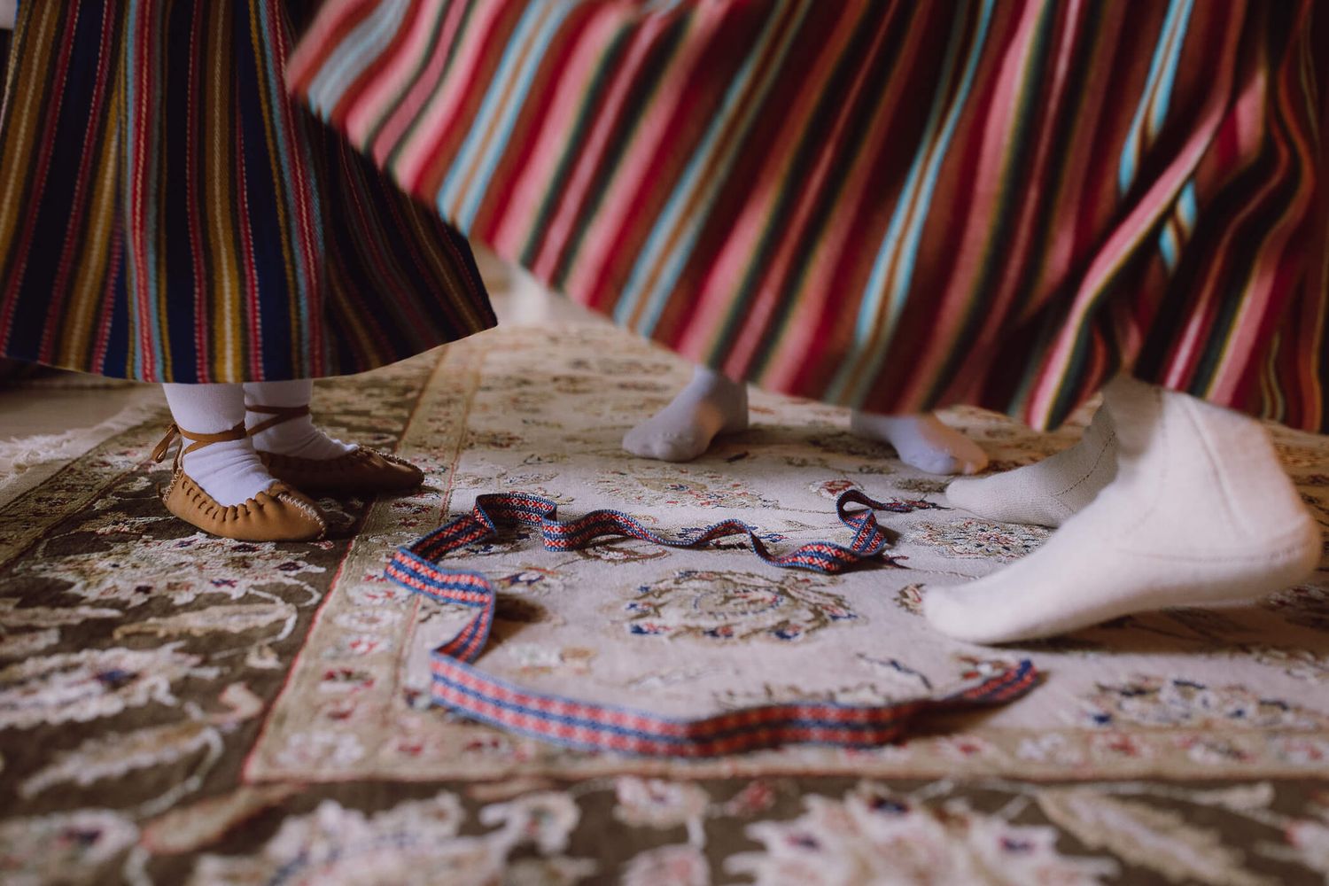 Close-up of feet in traditional folk costume shoes on ornate rug with colorful accessories scattered nearby.