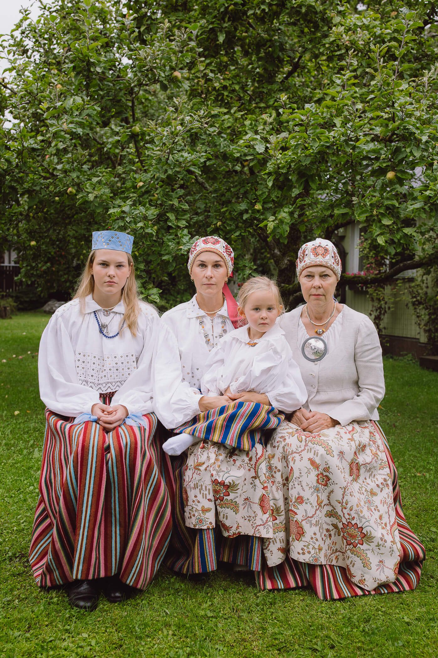 Four generations of women in traditional Estonian folk costumes sitting together outdoors near a green tree.