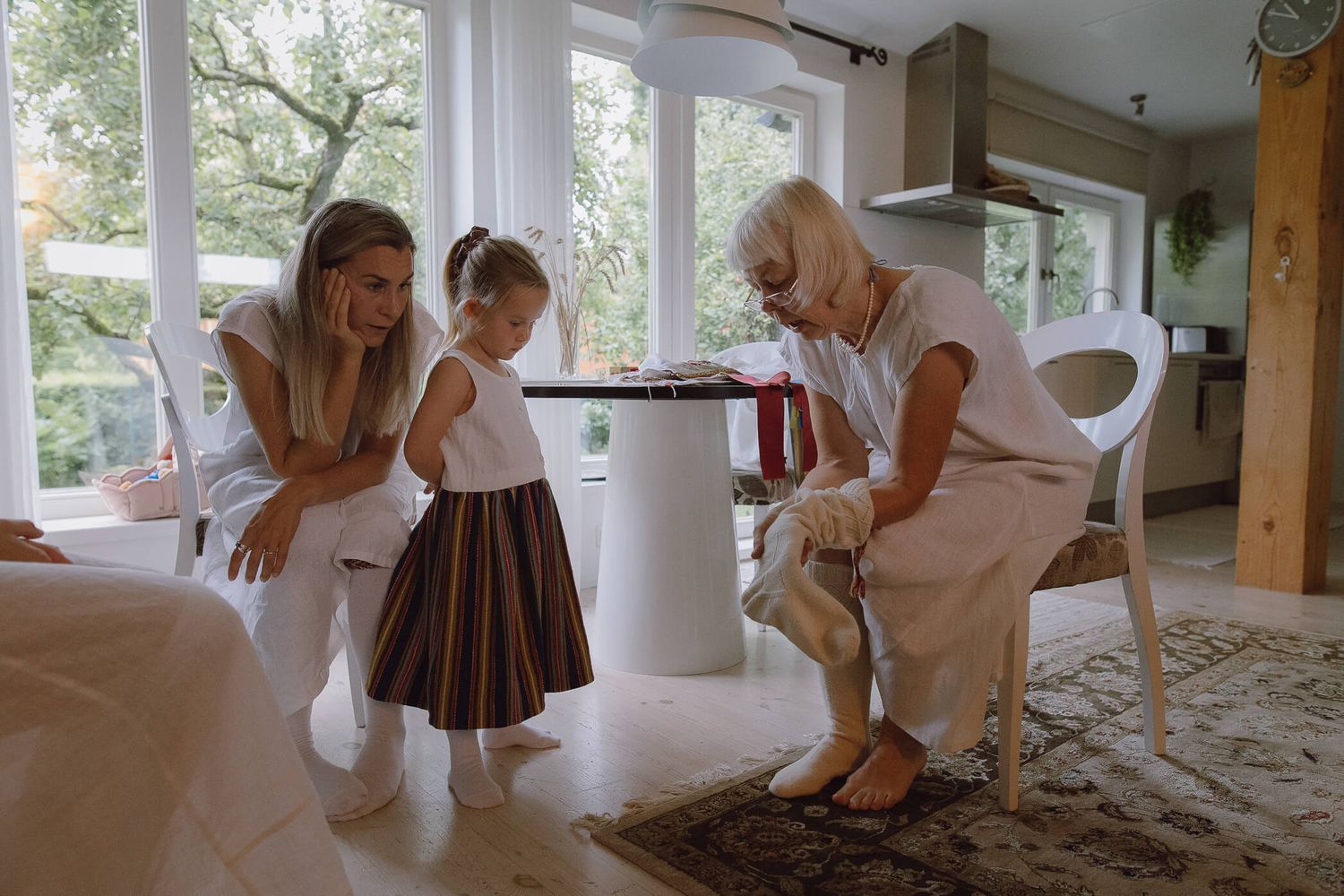 Grandmother helps young girl in traditional Estonian striped skirt while mother watches indoors.