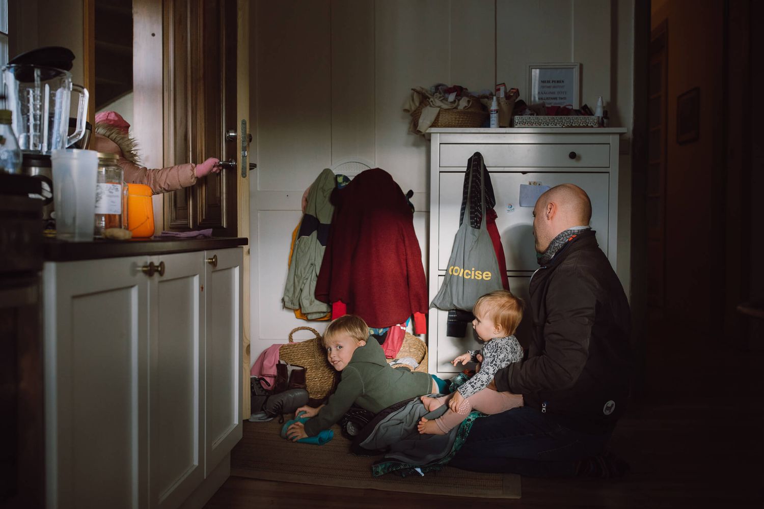 A family gathers in a dimly lit mudroom entryway, organizing shoes and coats near a white cabinet.