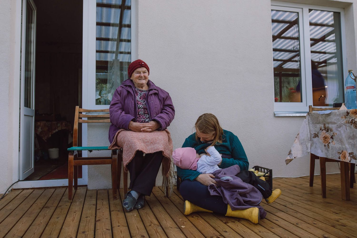 Two people sit on a wooden deck outside a house, one in a purple coat and another sitting lower.