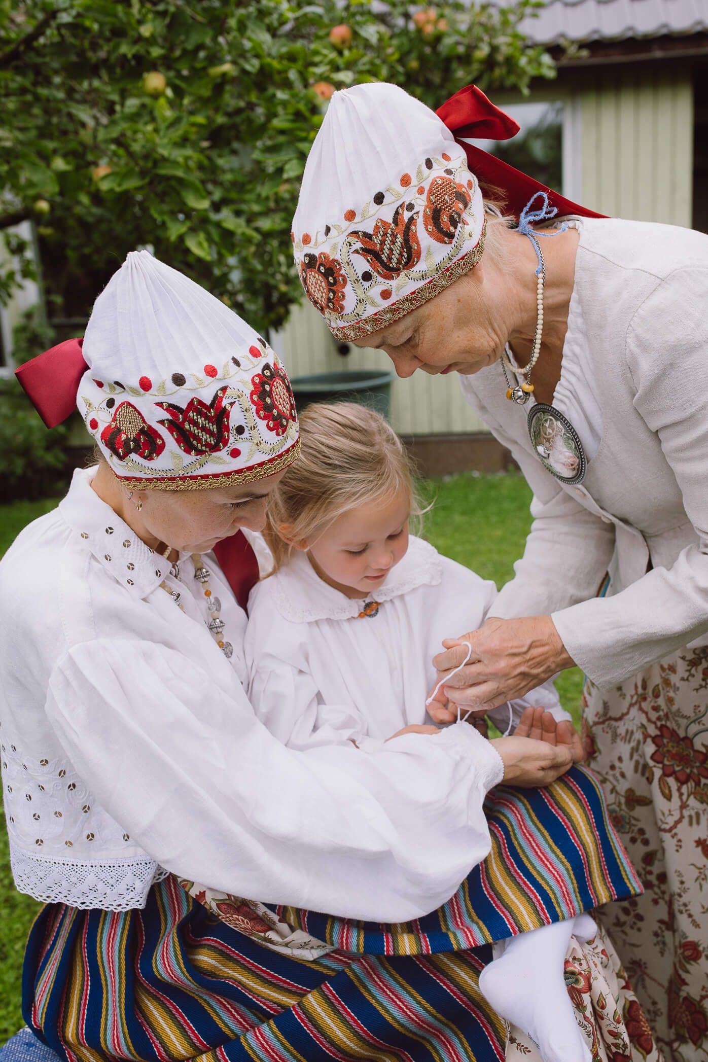 Women and child in traditional Scandinavian folk costumes with embroidered white caps outdoors.