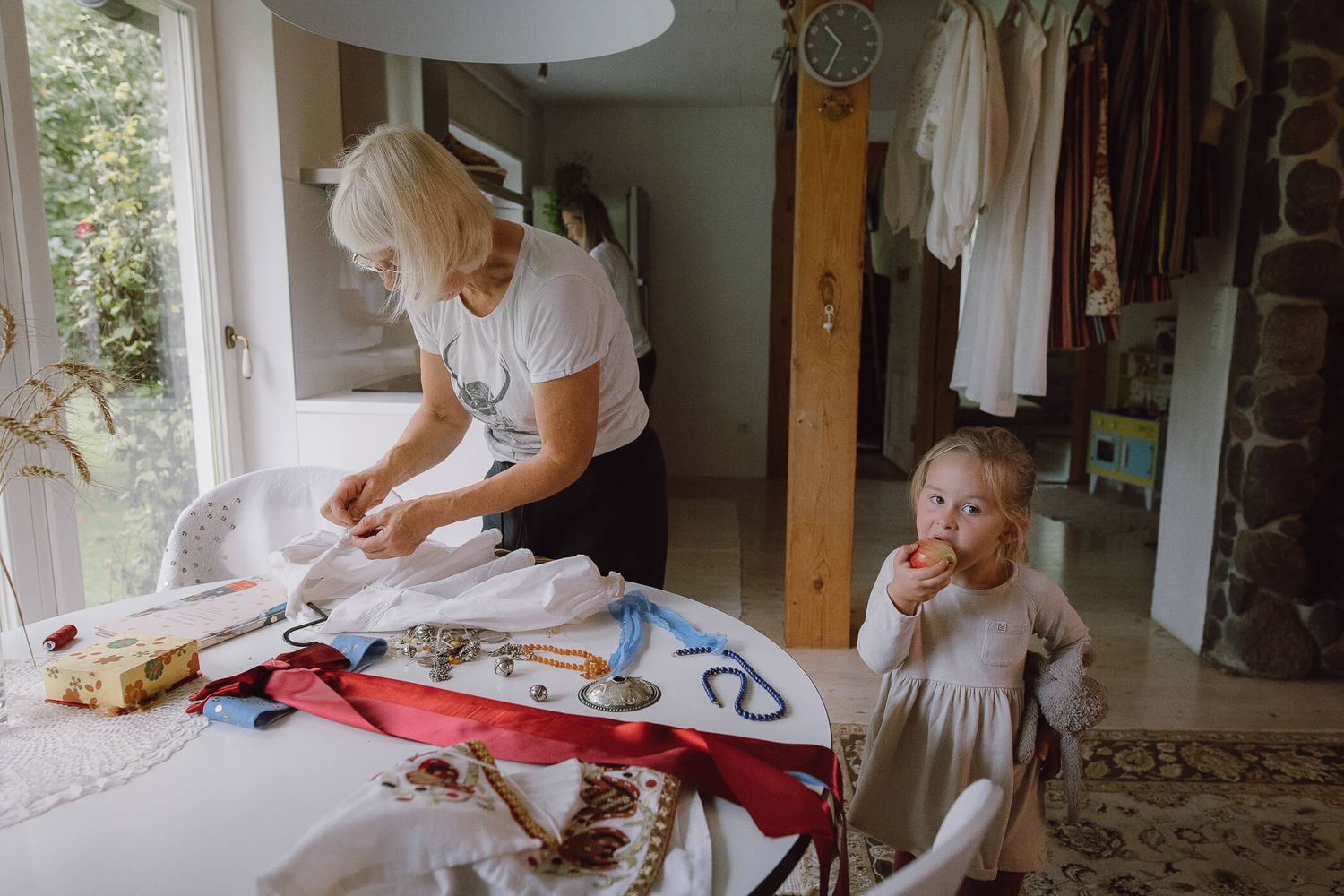 Grandmother irons clothes at a table while a young toddler girl stands nearby eating in a bright home interior.