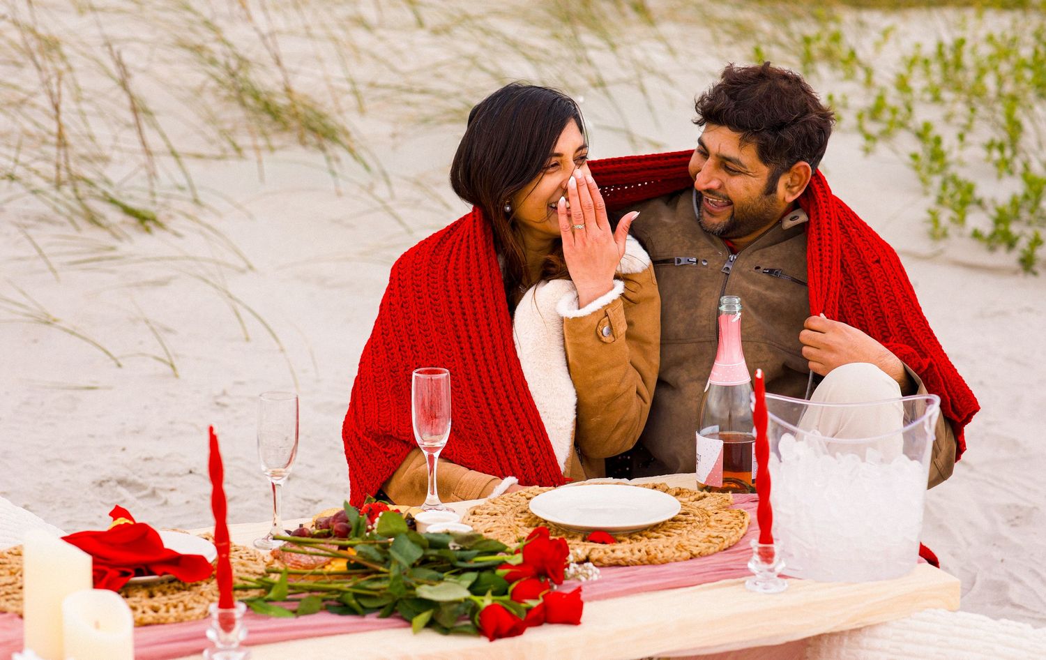 Couple wrapped in red blanket shares romantic beach picnic with roses and champagne.