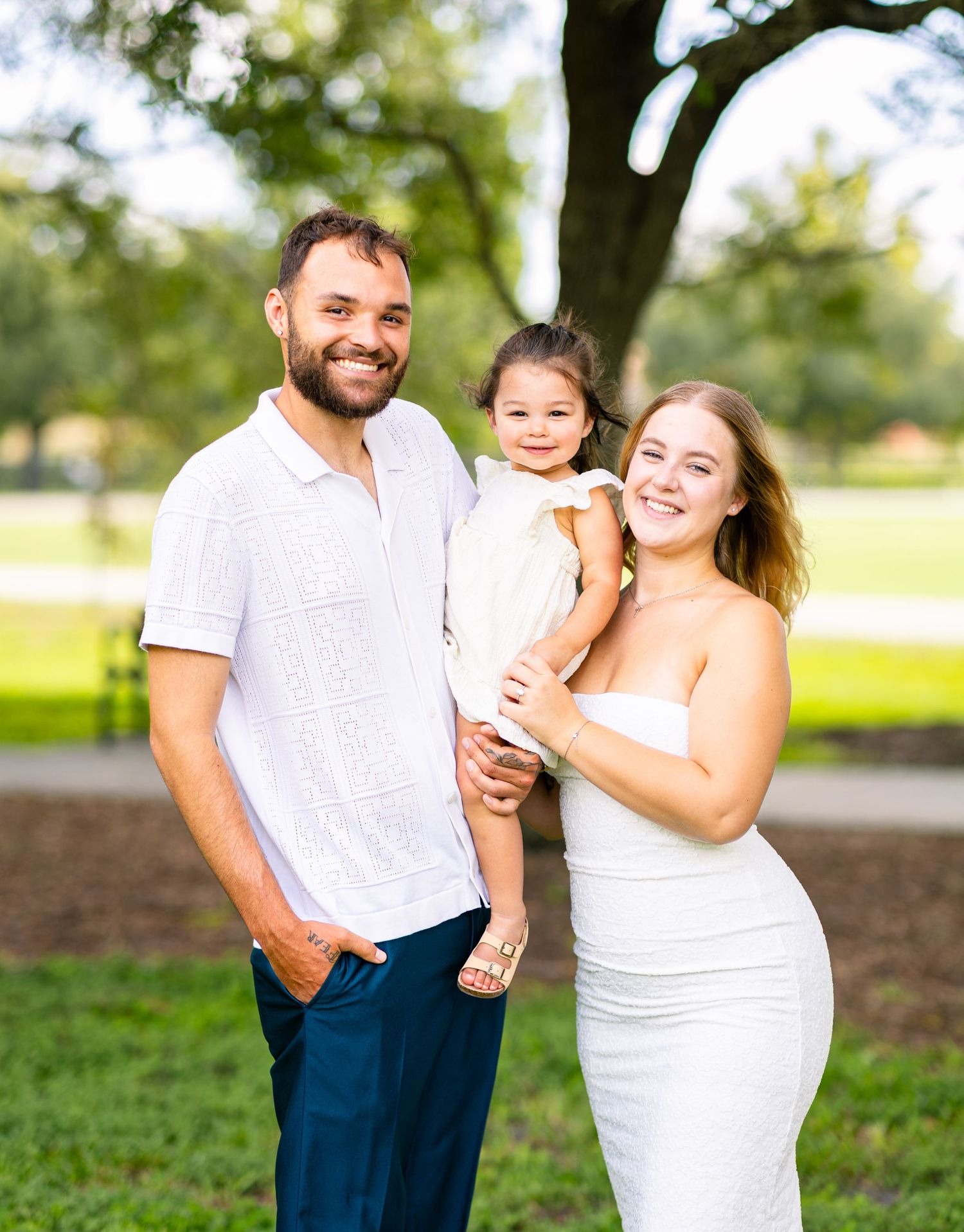 Smiling family of three in white outfits poses outdoors under a large oak tree in a sunny park in Orlando Florida.