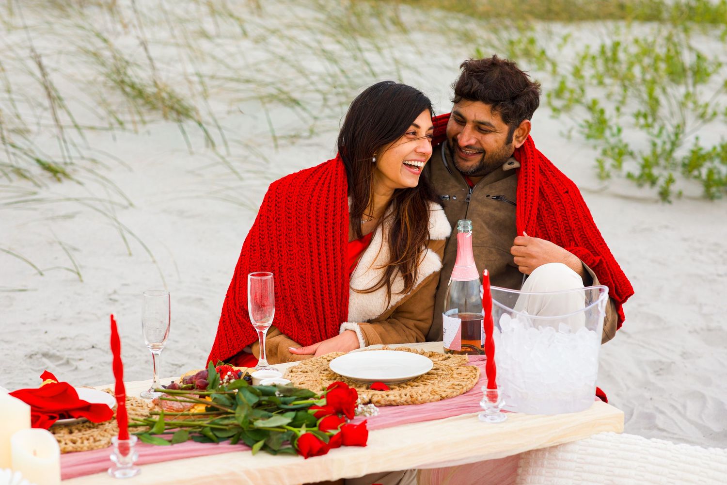 Happy couple shares romantic beach picnic under red blanket with roses and champagne.