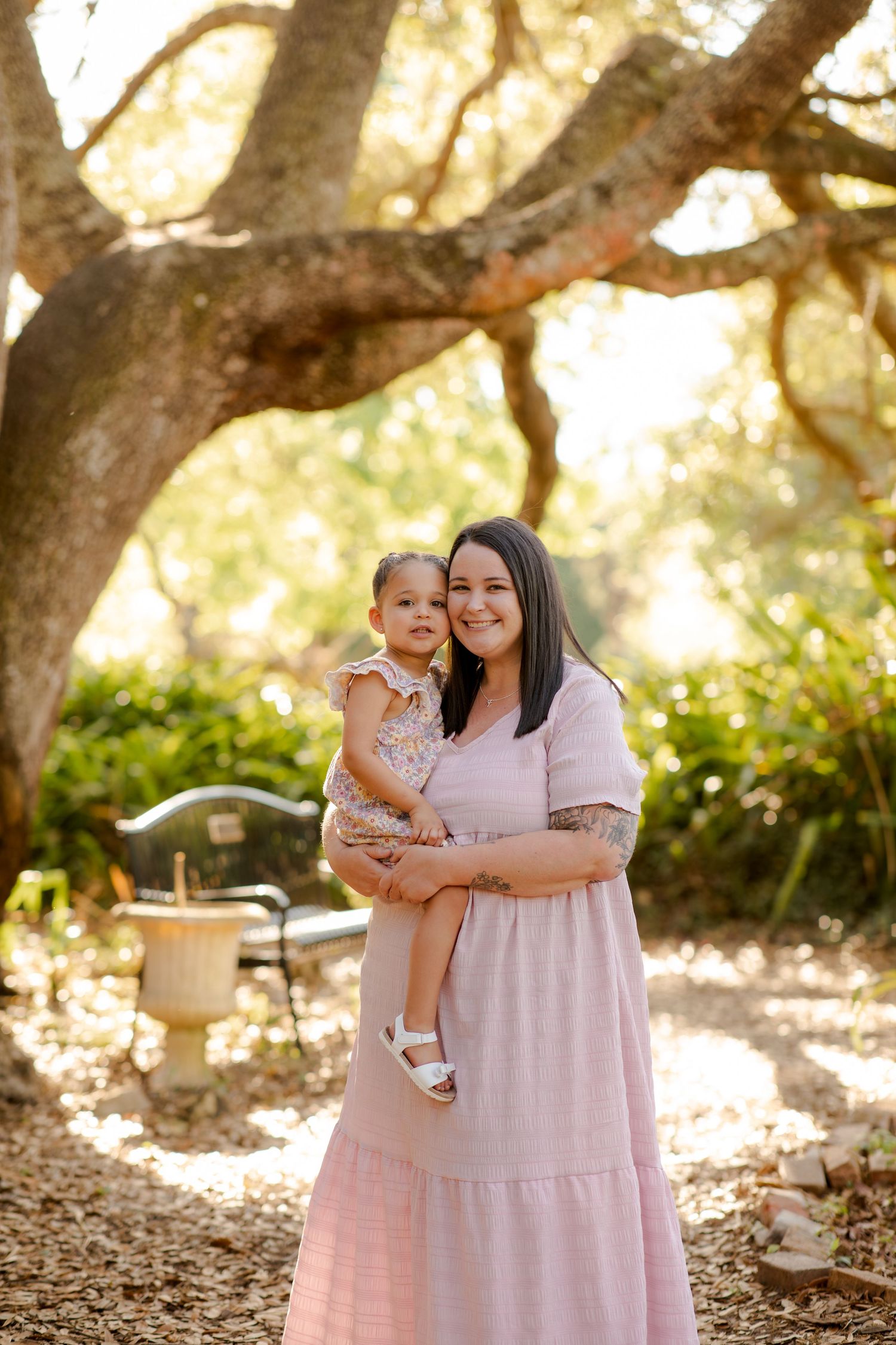 Smiling mother holds young toddler daughter in pink dress under large oak tree outdoors.