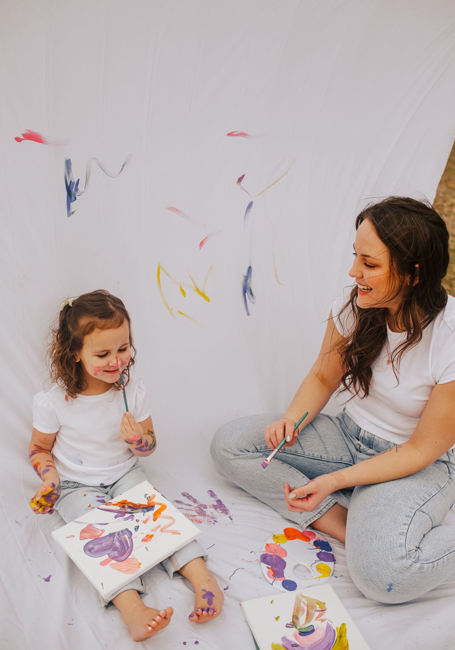 Mother and toddler daughter enjoy creative painting session on white drop cloth floor.