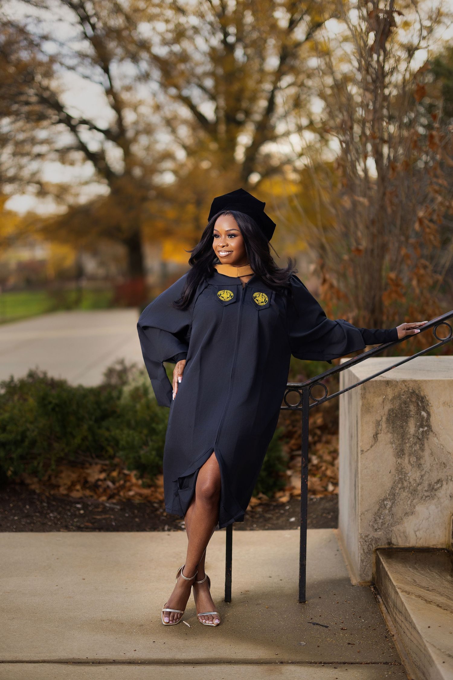 A person in a navy graduation gown poses on concrete steps surrounded by autumn trees and fallen leaves.