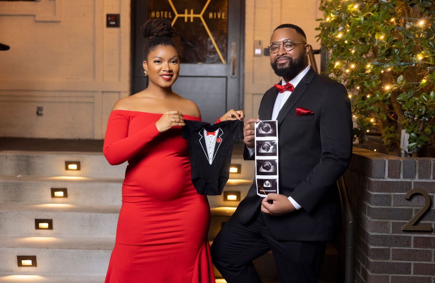 A couple poses together in formal attire in front of a decorated Christmas tree and elegant staircase.