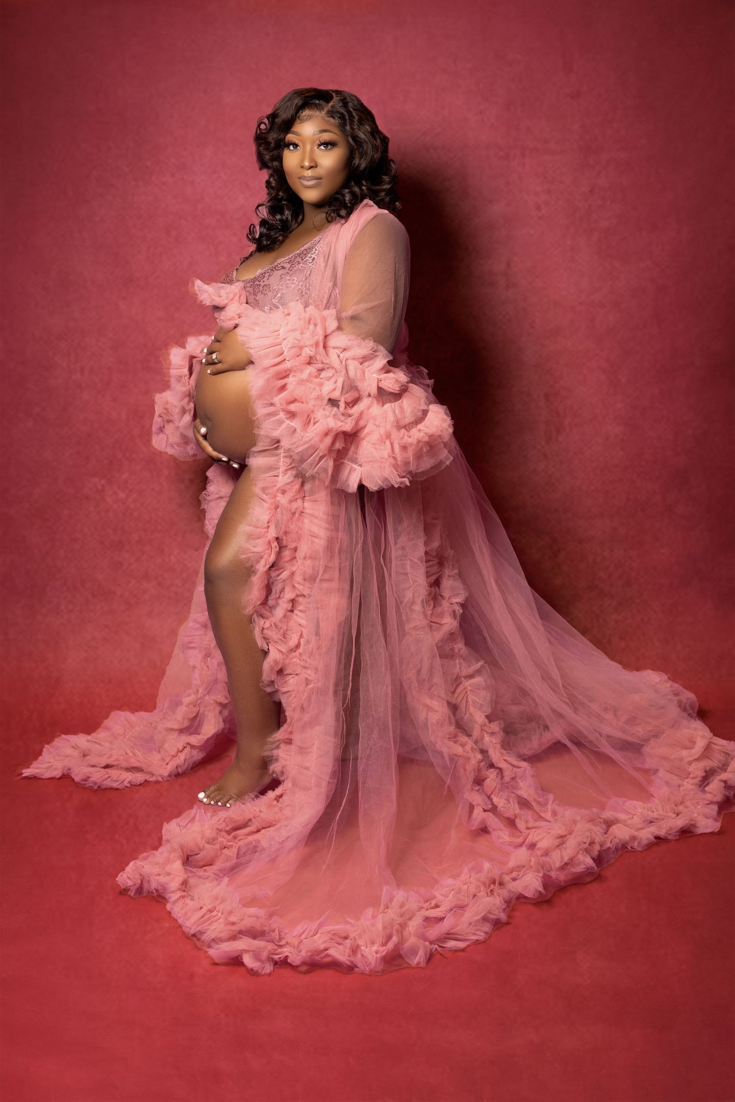 A glamorous maternity photoshoot features a flowing pink tulle gown against a deep red backdrop.