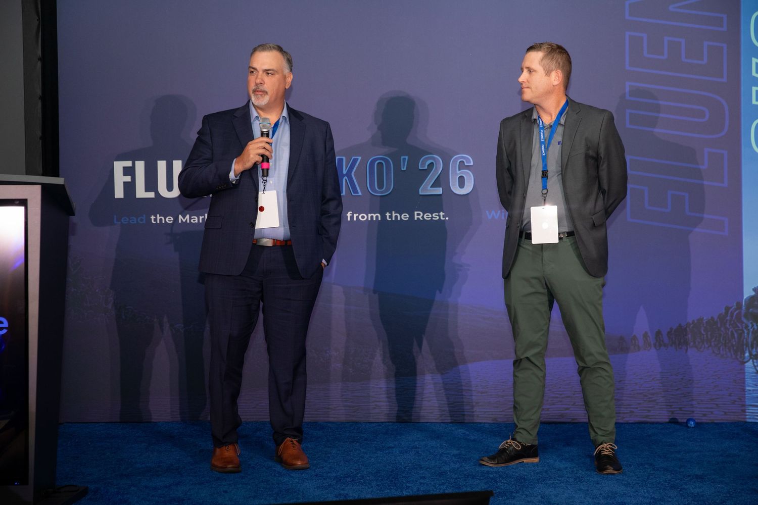 Two people gesture while speaking on stage at a business conference with blue lighting and signage.