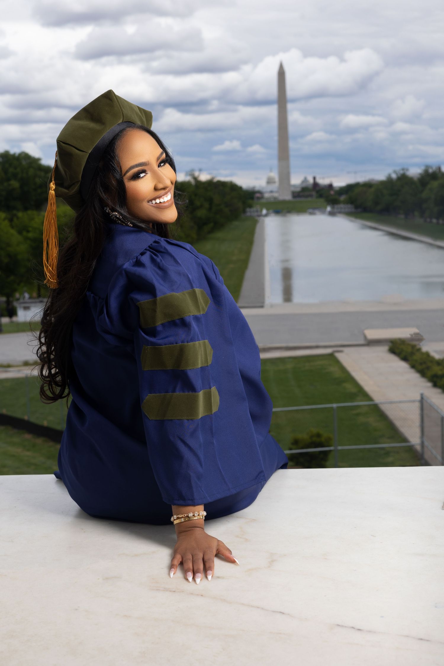 A graduate in academic regalia poses on a balcony with the Washington Monument visible in the background.