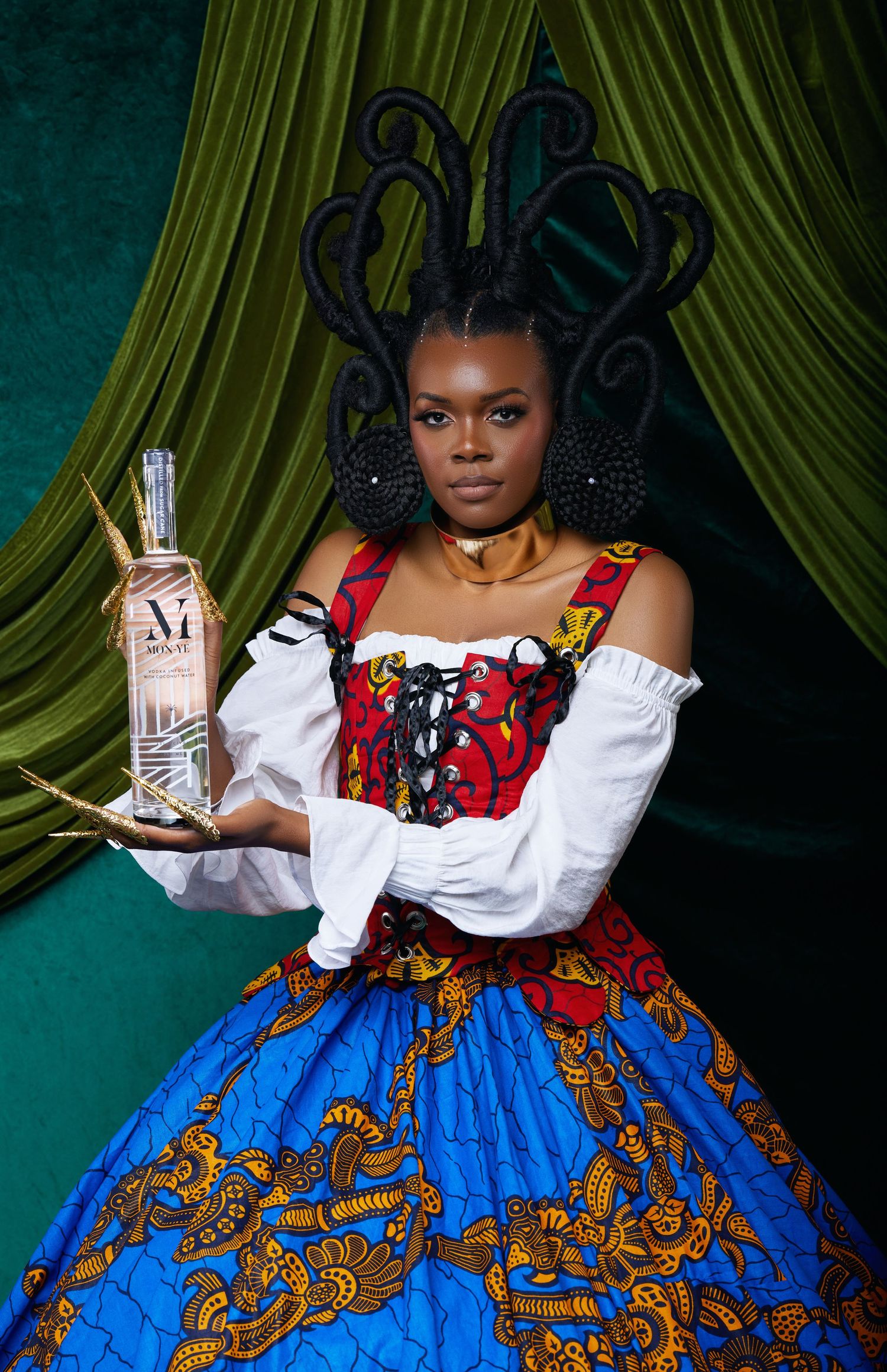 A model poses in a dramatic gown featuring a white bodice and vibrant blue African print skirt against a dark green backdrop.