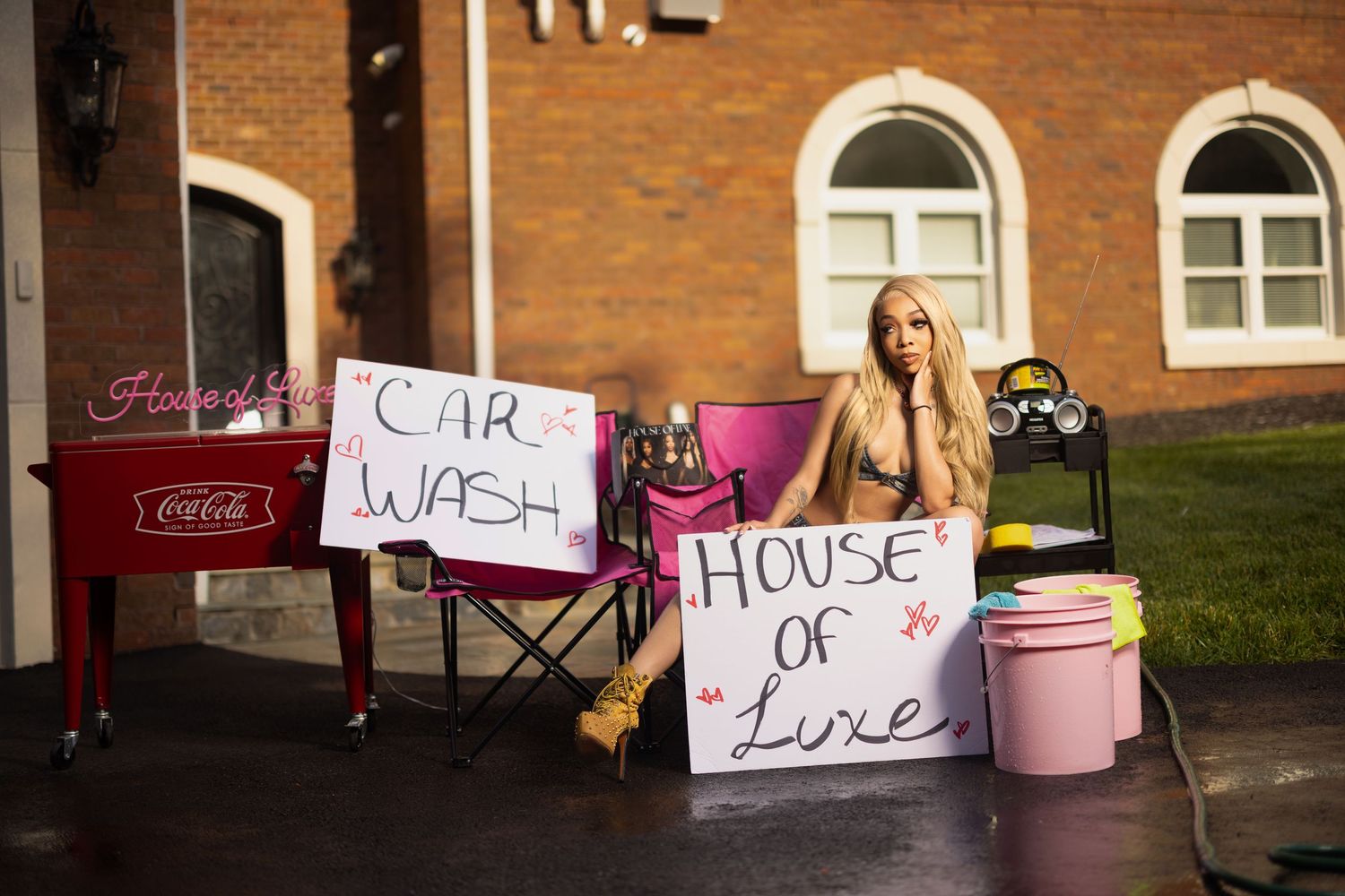 A makeshift car wash setup with handmade signs and cleaning supplies sits outside a brick building.