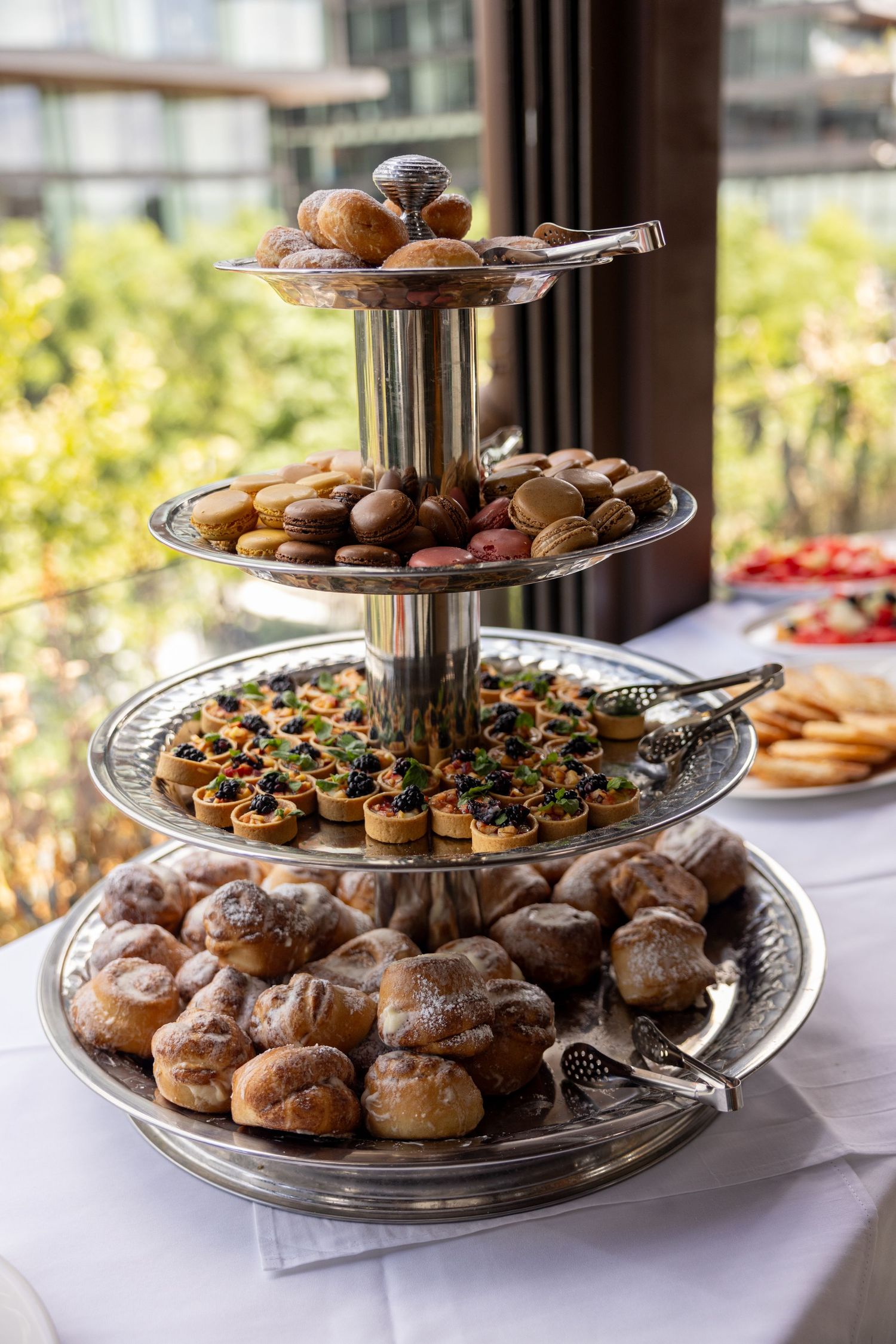 Elegant tiered dessert stand filled with assorted pastries, cookies and petit fours on a white tablecloth by a window.
