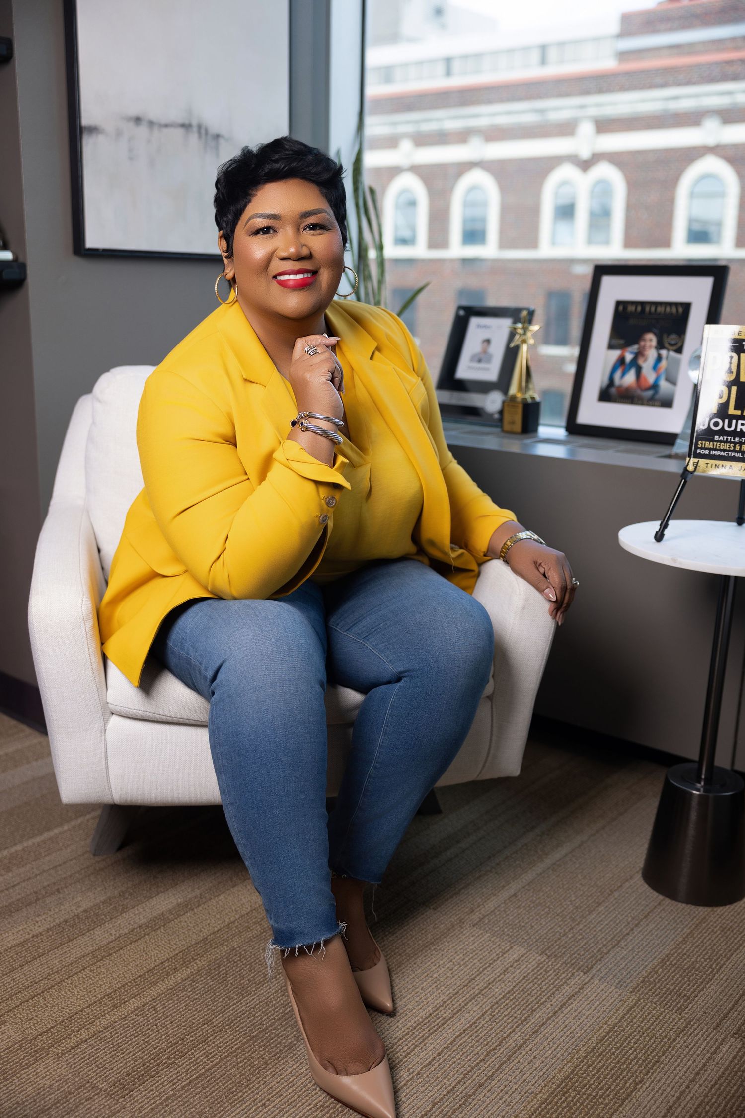 A person in a bright yellow blazer and jeans sits casually on a white chair in an office setting.