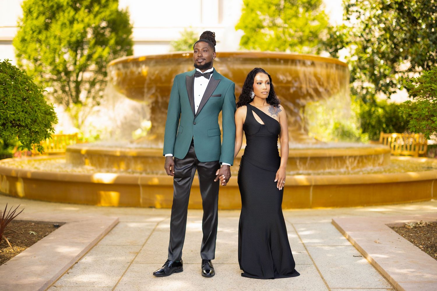A couple in formal attire walks along a garden path during golden hour as sunlight filters through the trees.