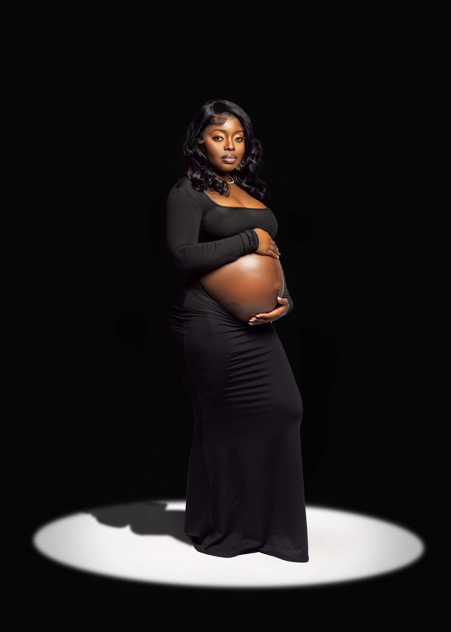 A pregnant woman in a black dress poses elegantly in a dramatic spotlight against a dark background.