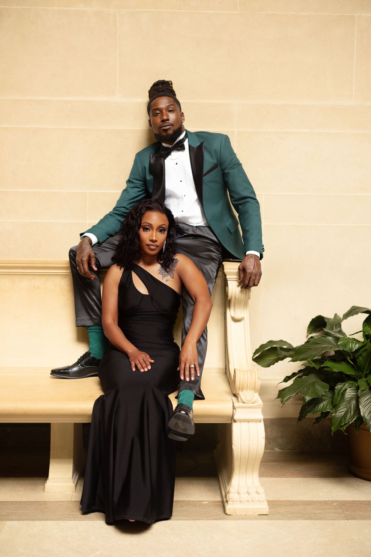 A couple in formal attire poses together on a bench, with the woman in a black dress and man in a green suit.