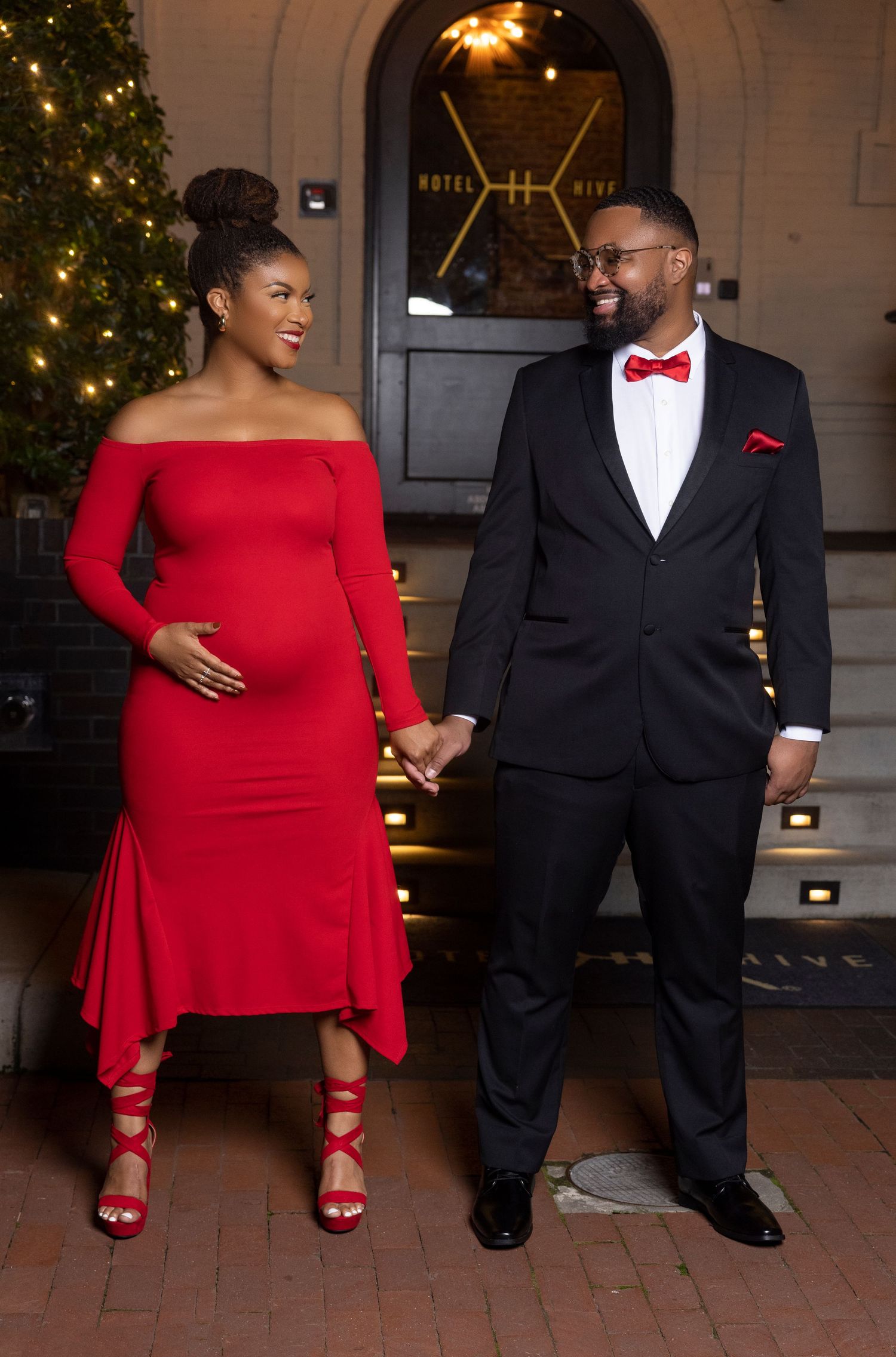 A couple in formal attire poses for holiday photos, with her in a red dress and him in a black tuxedo with bow tie.