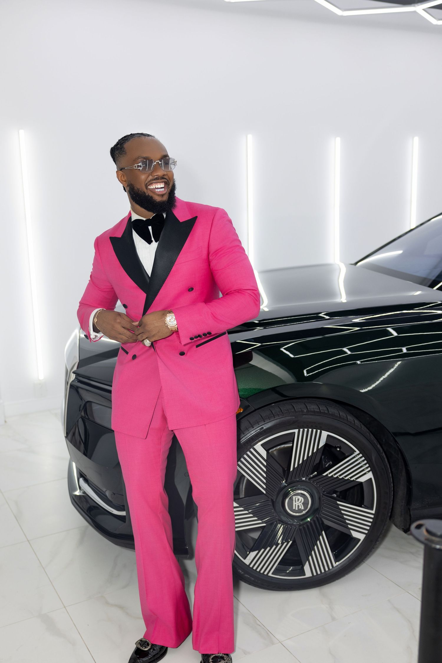 A person in a bright pink suit poses next to a sleek black luxury car in a white showroom.