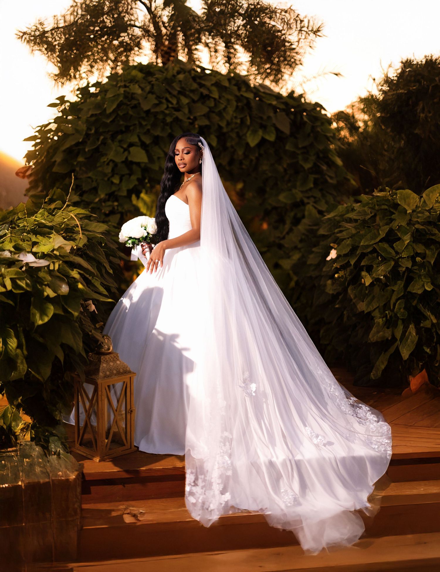 A bride in a flowing white wedding gown and long veil poses in a garden at sunset with lush greenery and lantern decor.