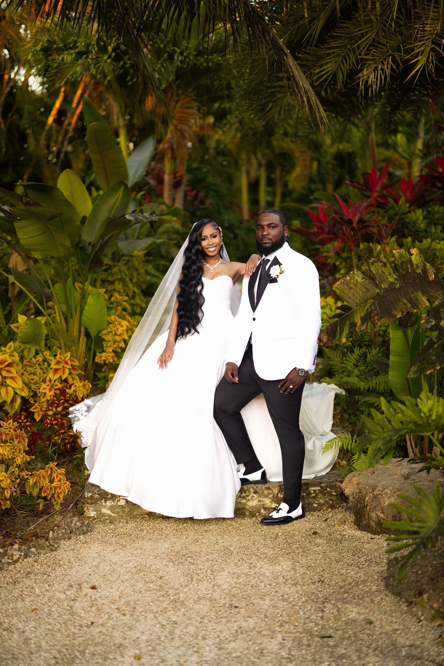 A couple in wedding attire poses together in a lush tropical garden setting with vibrant greenery and flowers.