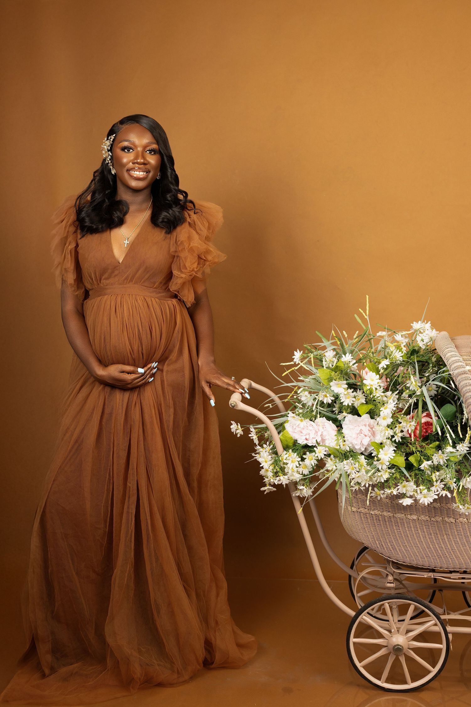 A maternity portrait with a brown flowing dress and decorative vintage carriage filled with white floral arrangements.