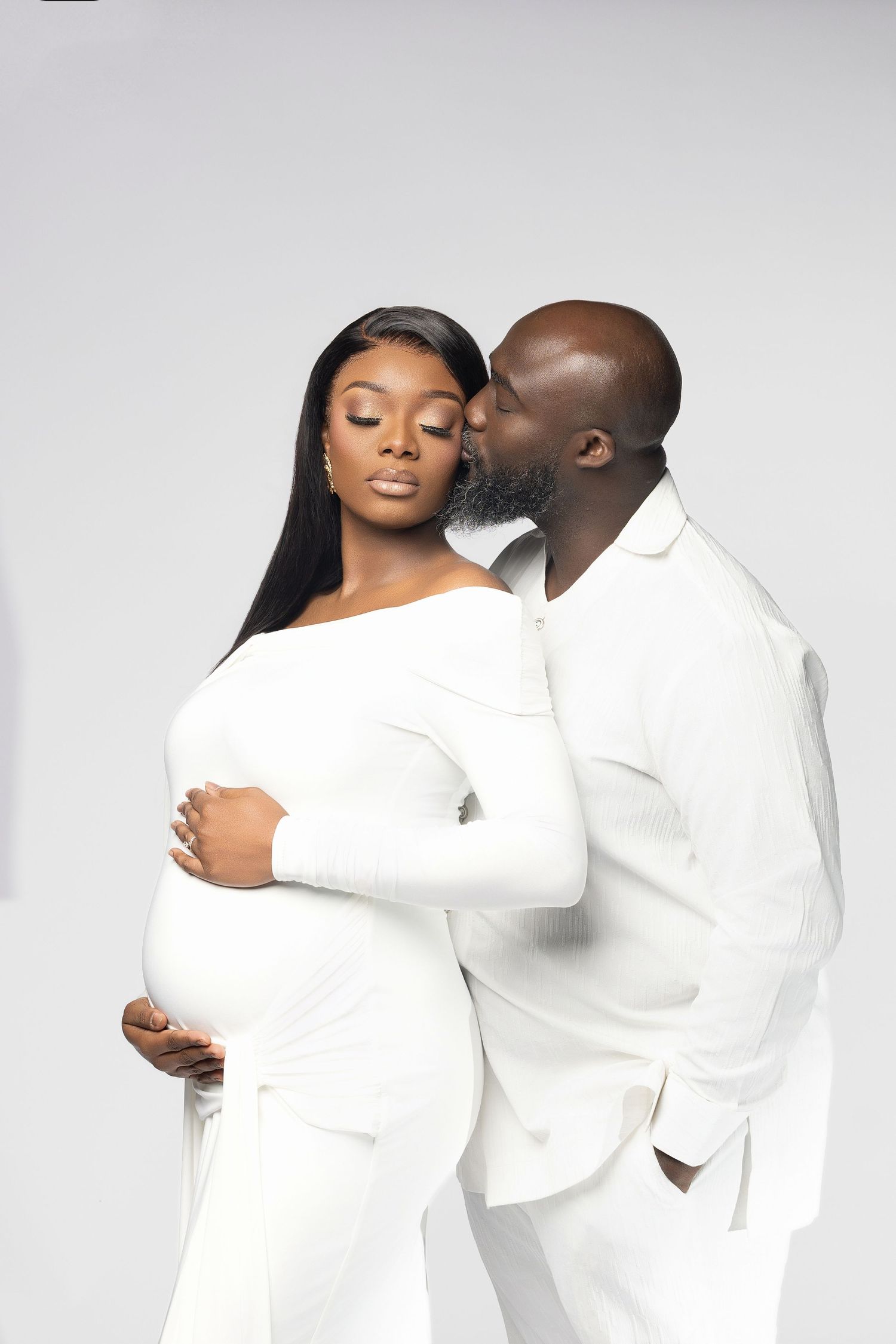 A romantic couple in matching white outfits embrace tenderly against a light gray studio background.