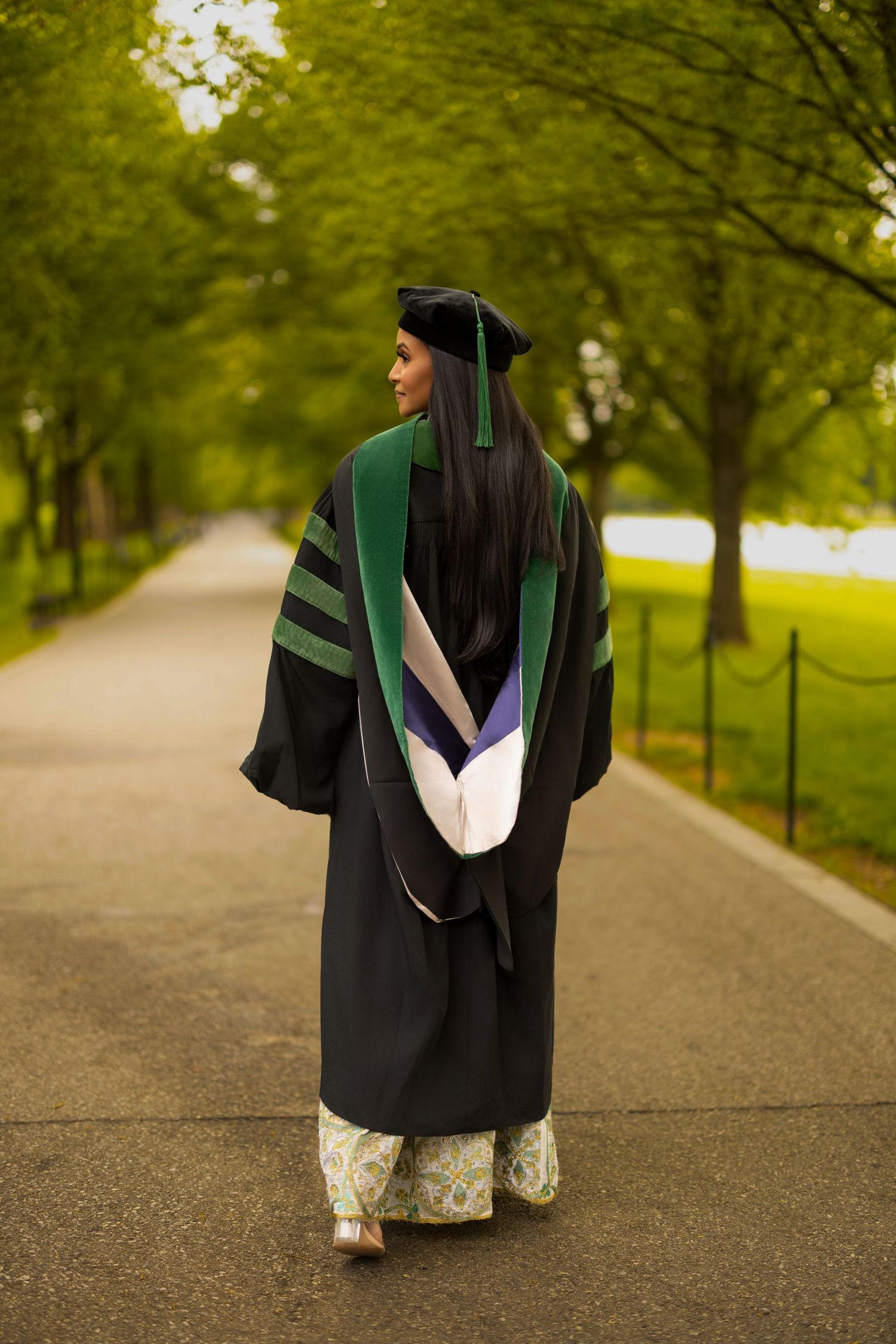 A graduate in a black cap and gown walks down a tree-lined path on a spring day.