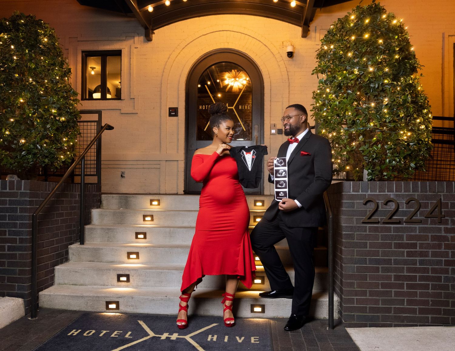 A couple in winter attire stands on illuminated steps near a decorated Christmas tree and brick building entrance.