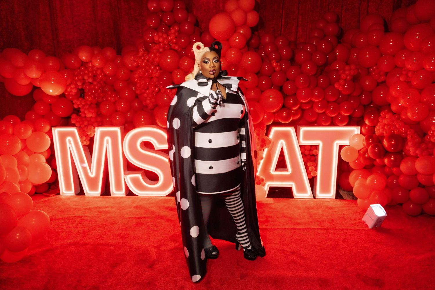 Dramatic red lighting illuminates figure in black and white striped costume against MS AT signage.